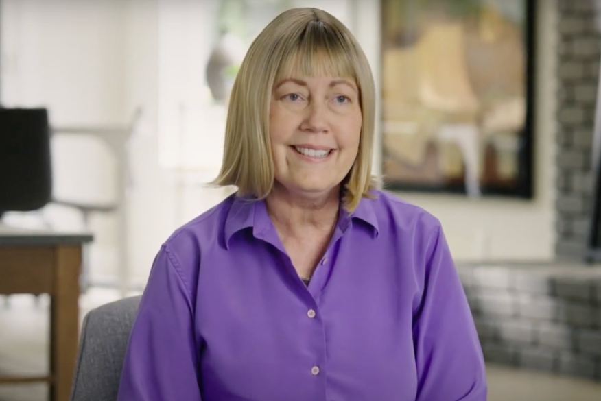 Esther smiles in a purple blouse, seated in her living room.