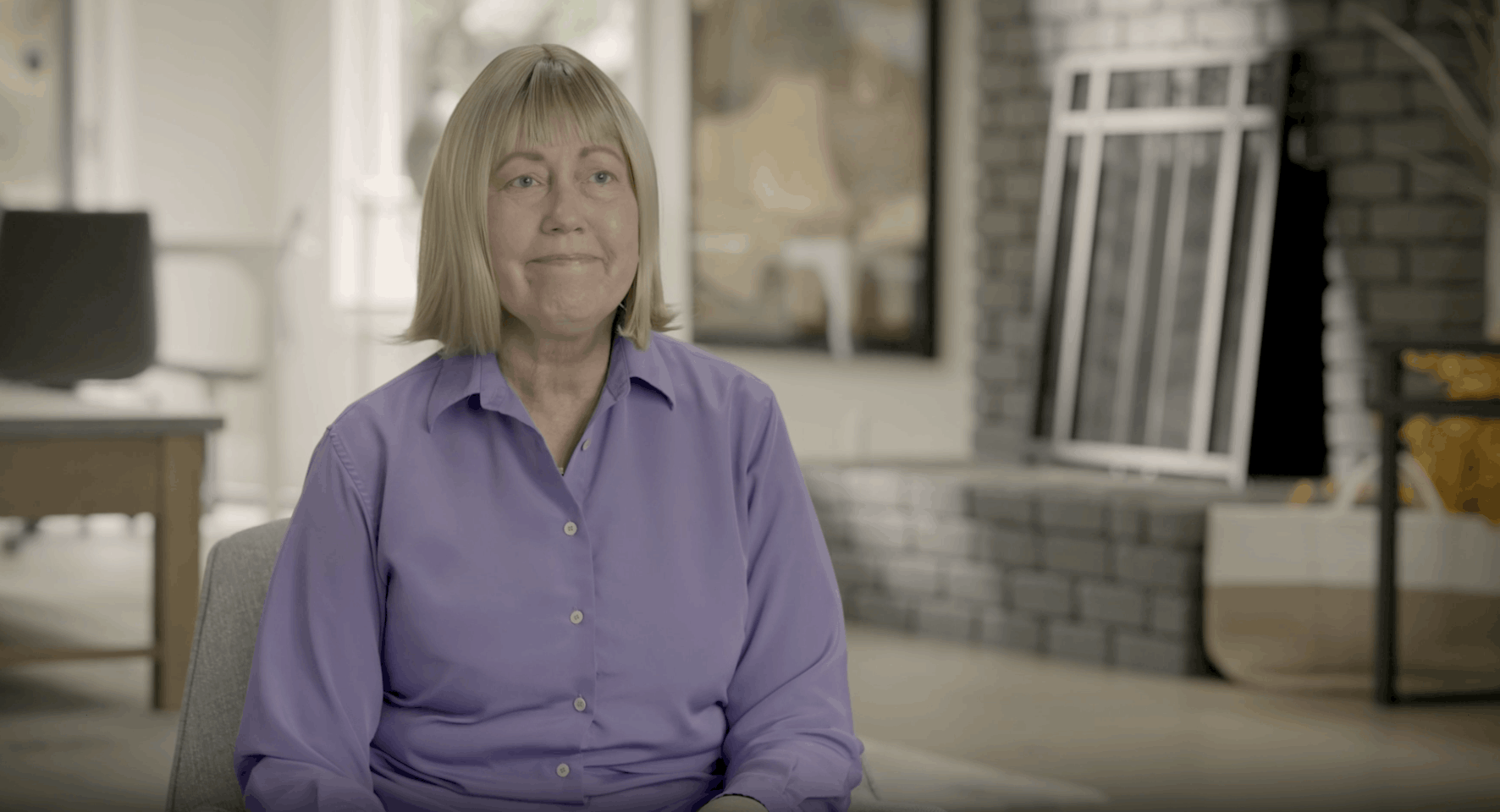Esther sits in her living room in a lilac shirt.