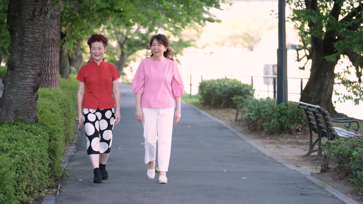Kei walks along a tree-lined path with a companion, both in bright clothing.