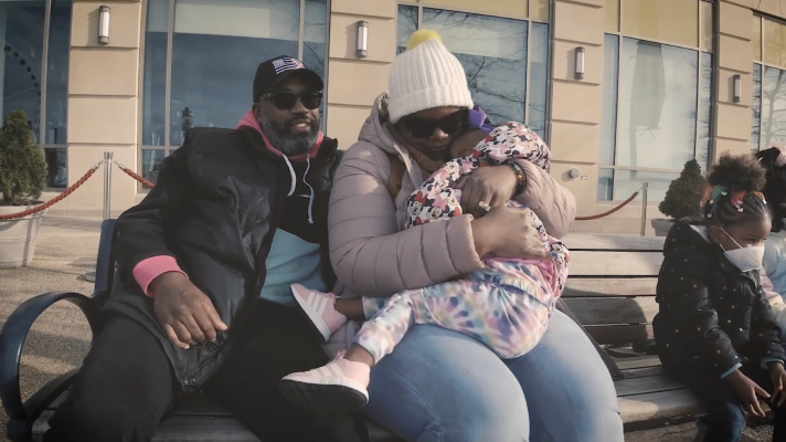 LaToya sits on a bench outdoors holding a child in a pink coat, with family beside her.