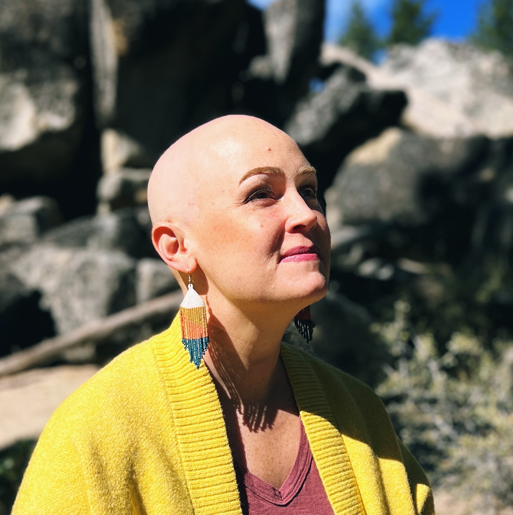 Lauren looks into the distance in a yellow cardigan, rocks and pine trees behind her.