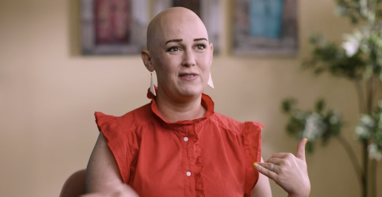 Lauren speaks, gesturing with one hand, in a coral blouse.