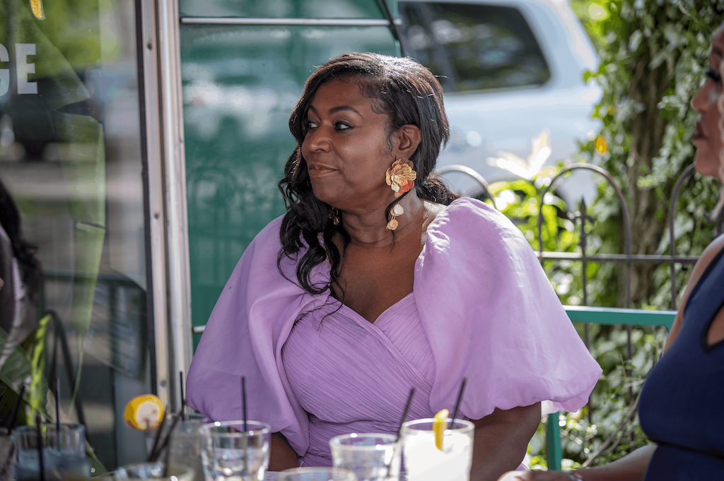 Vanessa sits at an outdoor cafe table in a lilac blouse and gold earrings.