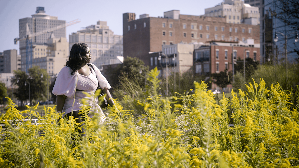 Vanessa stands among tall yellow wildflowers with a city skyline behind her.