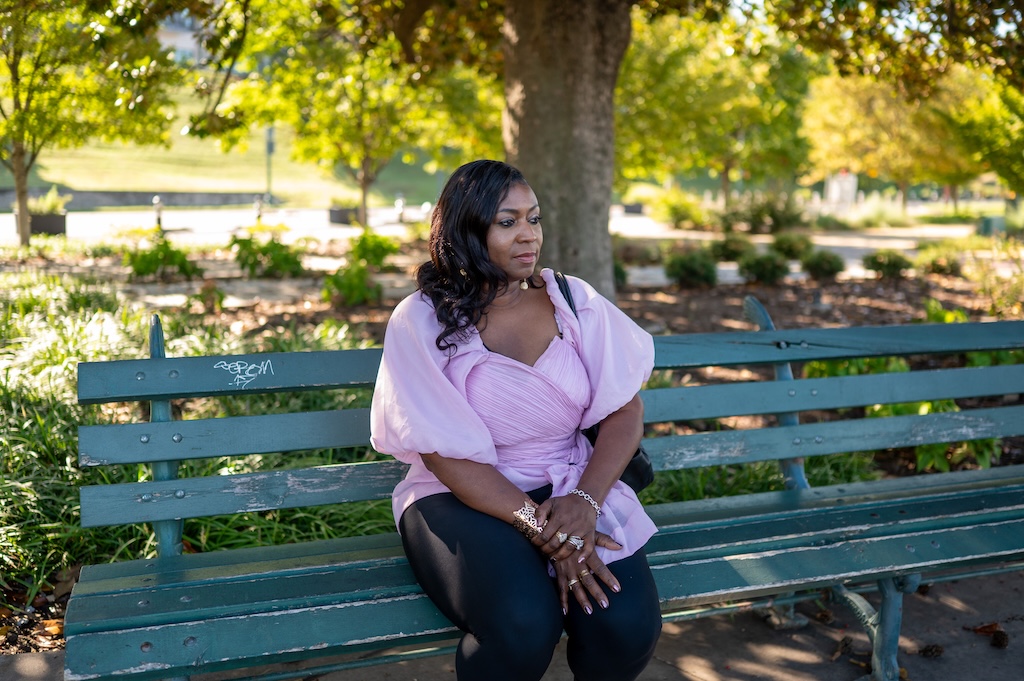 Vanessa sits on a park bench in a lilac blouse on a sunny day.
