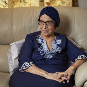 Joella sits in a cream leather armchair wearing a navy headwrap, glasses and a navy top with pale blue embroidery, her hands folded in her lap.