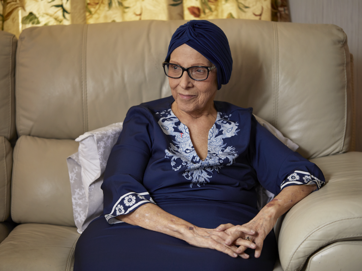 Joella sits in a cream leather armchair wearing a navy headwrap, glasses and a navy top with pale blue embroidery, her hands folded in her lap.