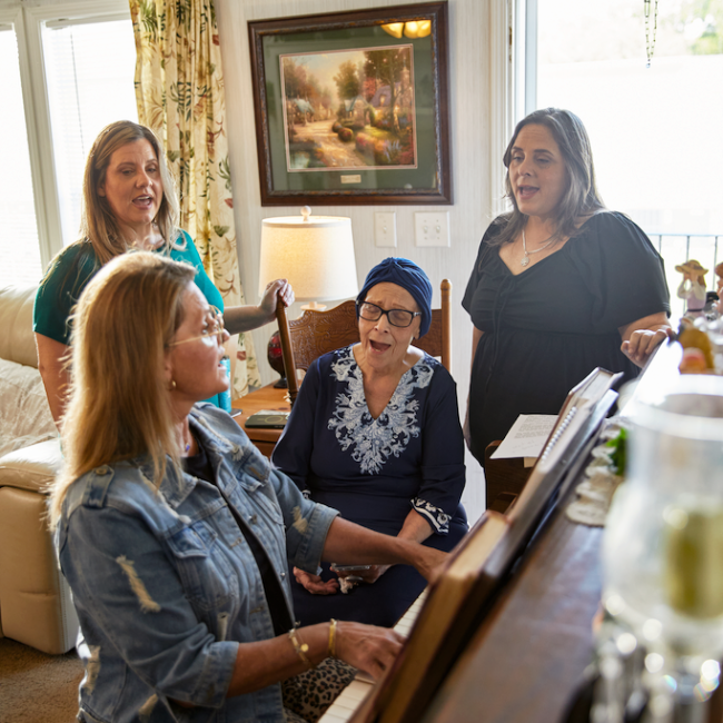 Joella sings by an upright piano in her living room with her family singing and playing around her.