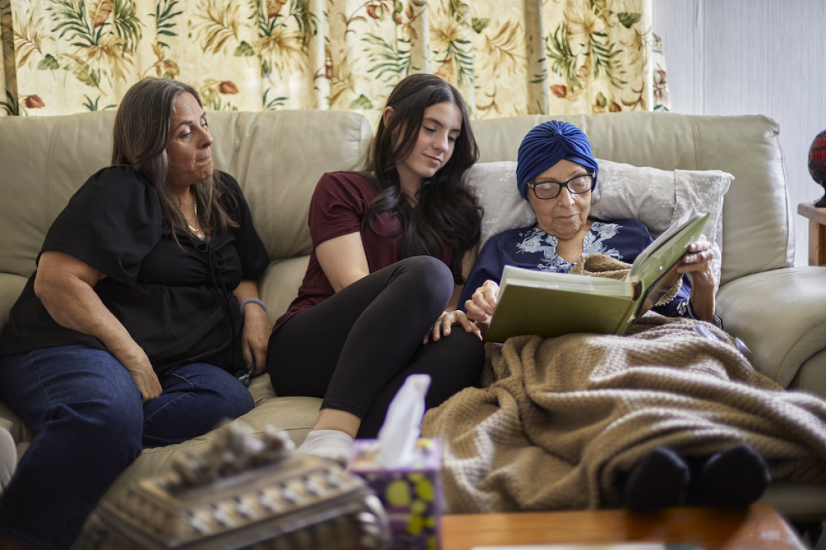 Joella looks through an open photo album on a sofa with her daughter and granddaughter beside her.