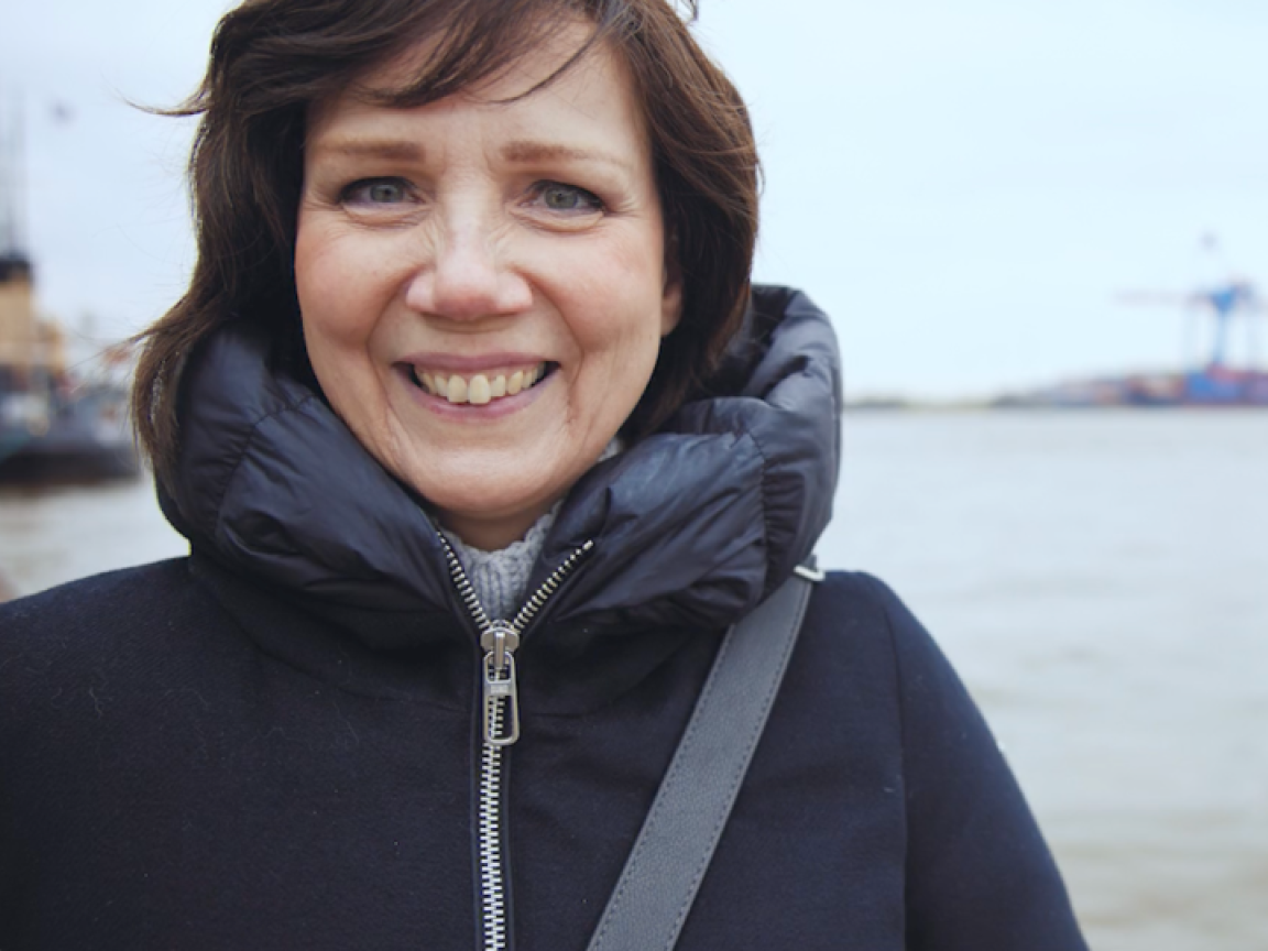 Katja smiles in a dark padded jacket with a harbor behind her.