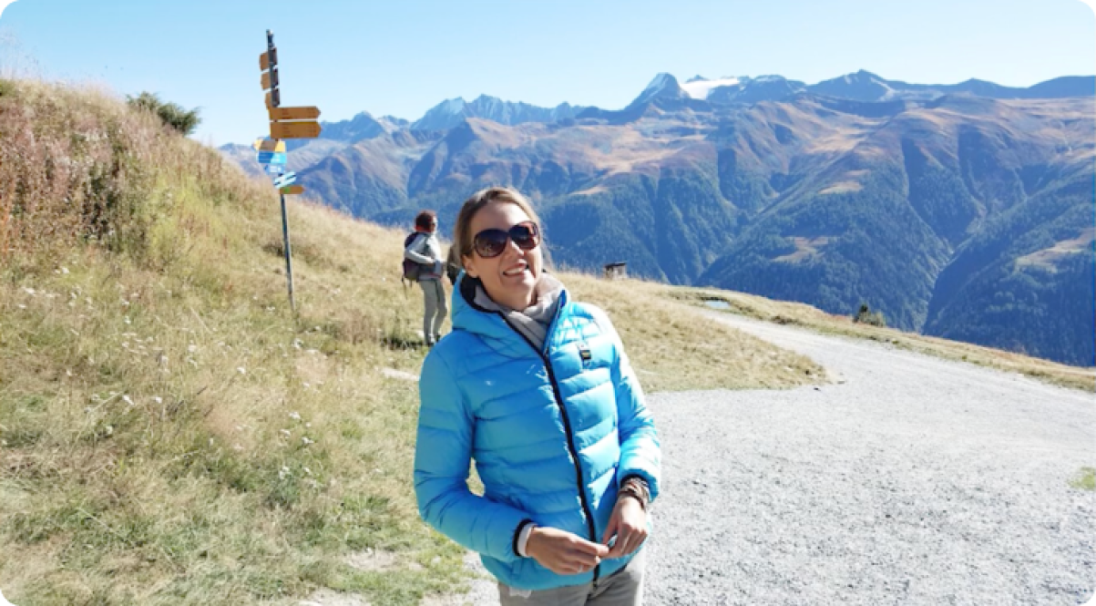 Katja stands on a mountain path in a blue jacket with peaks behind her.