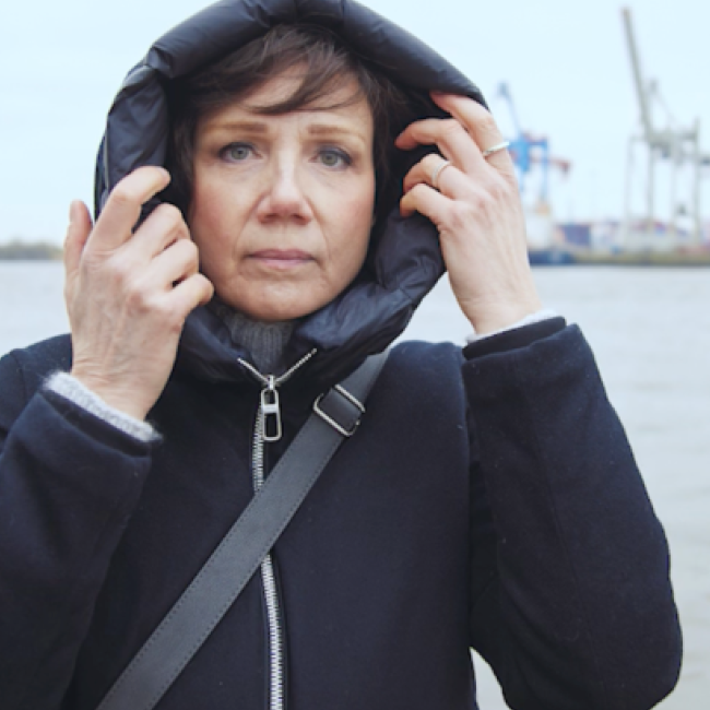 Katja pulls up the hood of her coat at a harbour, cranes behind her.