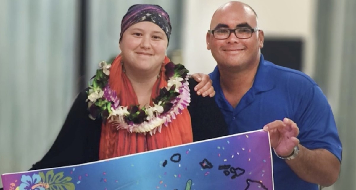 Melissa wears a flower lei and holds a painted board, with a man beside her.
