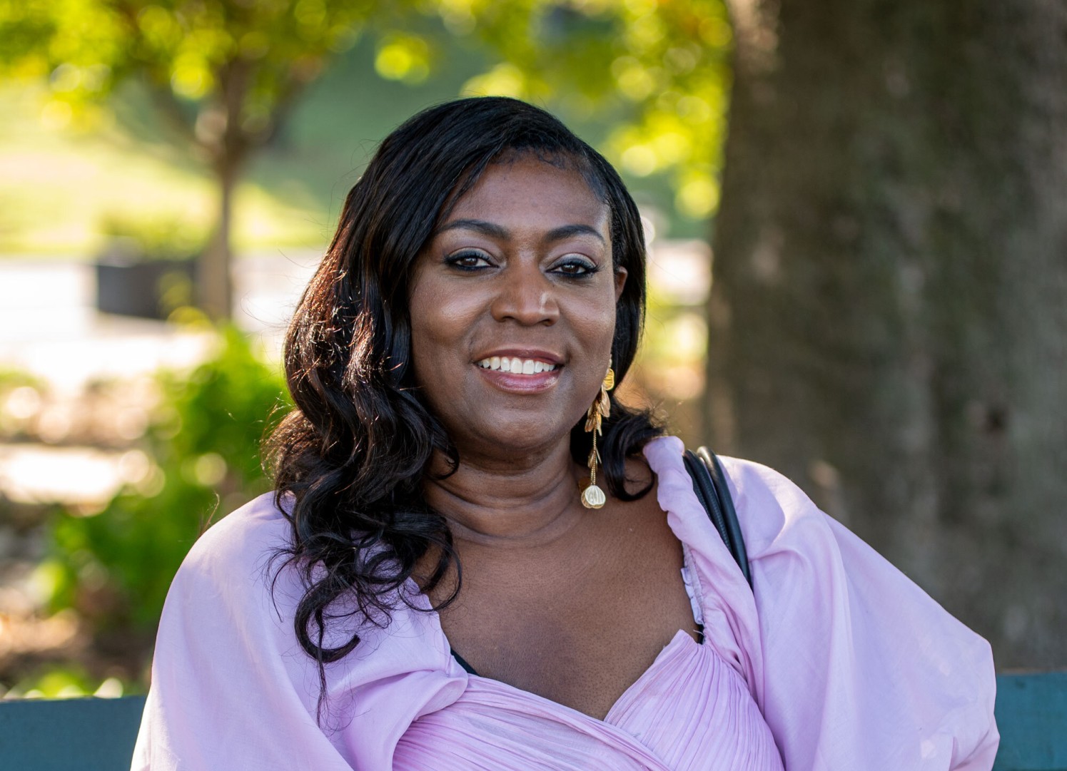Vanessa smiles outdoors in a lilac blouse, trees behind her.